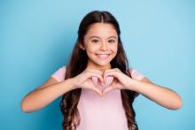 A young girl wears a pink shirt and makes a heart shape with her hands