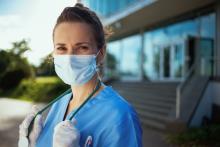A doctor wearing a mask stands outside of a hospital, looking determined