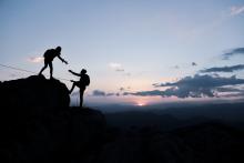 Two silhouettes are seen climbing a mountain; one reaches out a hand to pull the other up