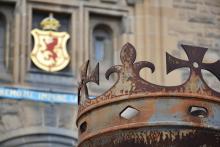 A crown in front of the gate to Edinburgh Castle, with the Royal Stuart coat of arms in the background