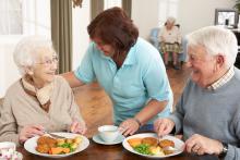 A care worker attends to a senior couple eating a meal at a care facility