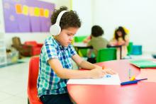 A young autistic child sits at a table in a classroom, wearing noise-canceling headphones and coloring.