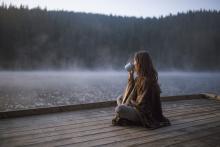 A woman sits on a dock overlooking the water, wrapped in a blanket and drinking from a mug. 