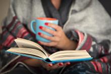 A woman wrapped in a blanket holds a book and a mug.