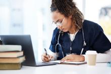 A doctor writes in a notebook while working on a computer.