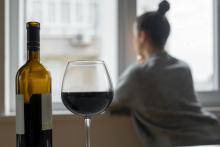 A woman sits on a couch with a bottle of wine and a poured glass behind her.