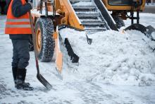 A person holding a shovel stands next to a snow plow clearing snow.