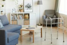 A living room in a long-term care facility, with a small couch, a chair, a lamp, a bookshelf, and a coffee table, with a walker in the foreground.