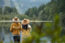 Two people wearing traditional Indigenous clothing stand side by side looking out on a lake.