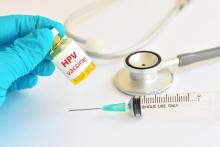 Gloved hand holds vial of HPV vaccine, syringe and stethoscope on table
