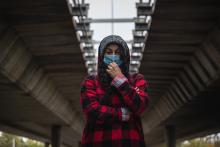 A young homeless person wearing a mask stands alone under a bridge 