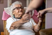 Female carer supporting old woman to stand up from an armchair at care home